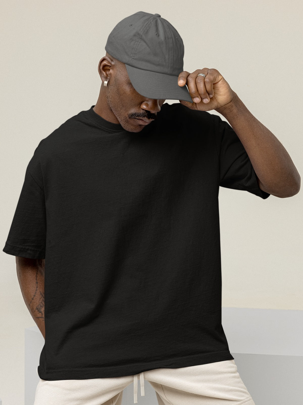 In this captivating image, a stylish black gentleman exudes confidence and charisma. He wears our Premium Heavyweight Black Oversized Tee with effortless coolness. A fashionable baseball cap adds a touch of urban flair to his look, and his downward gaze exudes a sense of contemplation. The tee, in classic black, drapes comfortably, embodying both comfort and a bold, timeless style that perfectly complements his individuality.