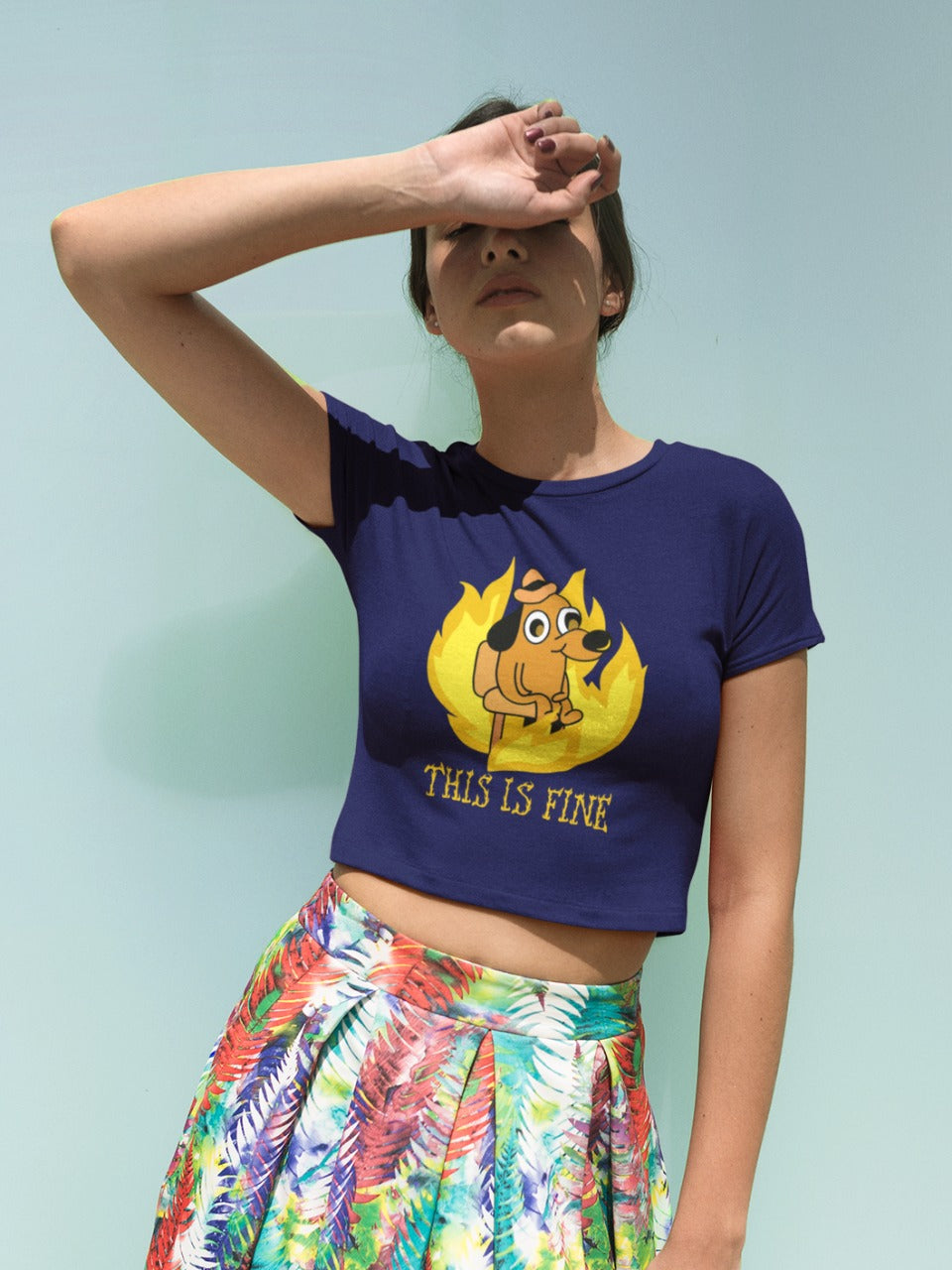 girl covering her face from the sun, wearing a colourful skirt and a navy blue crop top with small dog sitting around a fire printed on it, "this is fine" in boring letters is printed below the dog, relatable sarcastic memes