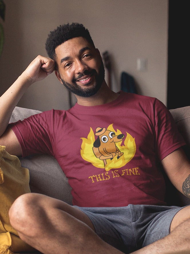 man sitting cross legged on a couch wearing shorts and a maroon tshirt with a small dog sitting around a fire printed on it, "this is fine" in burning letter is printed below the dog, relatable sarcastic funny memes