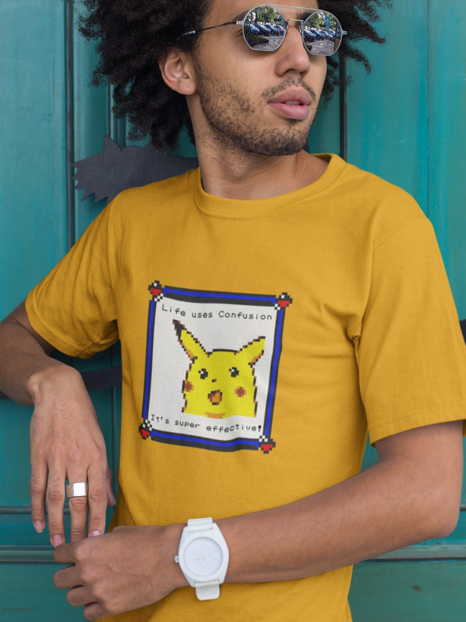 man with curly hair wearing sunglasses in front of a green door wearing a yellow tshirt with a console style photo of confused pikachu printed on it with message of "Life uses Confusion It's super effective", relatable Pokemon memes