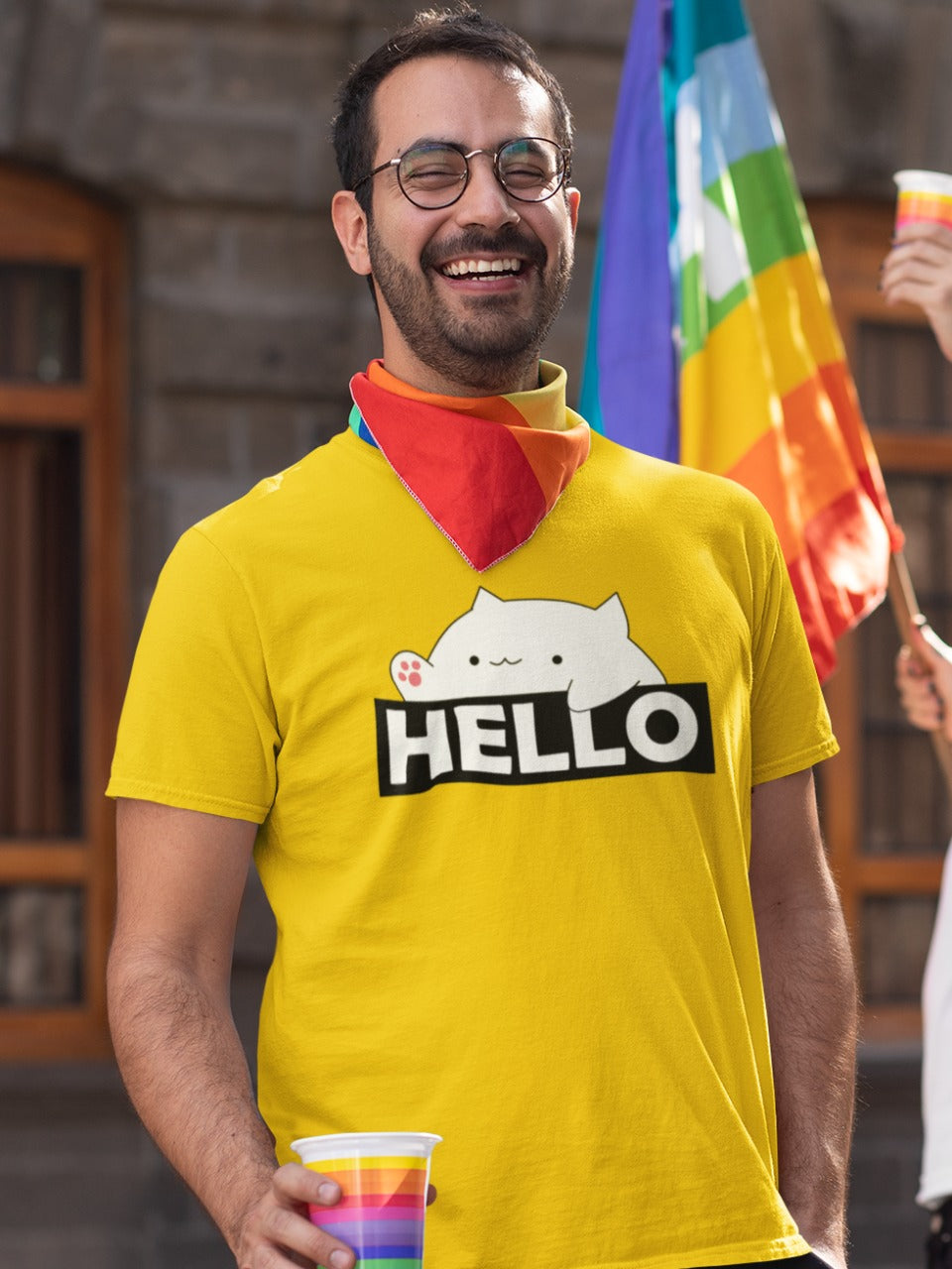 smiling man with spectacles wearing yellow tshirt with bongo cat saying hello written on it. The man has a rainbow pride cup in his hand and a rainbow lgbtq bandana. There is a rainbow pride flag in the background