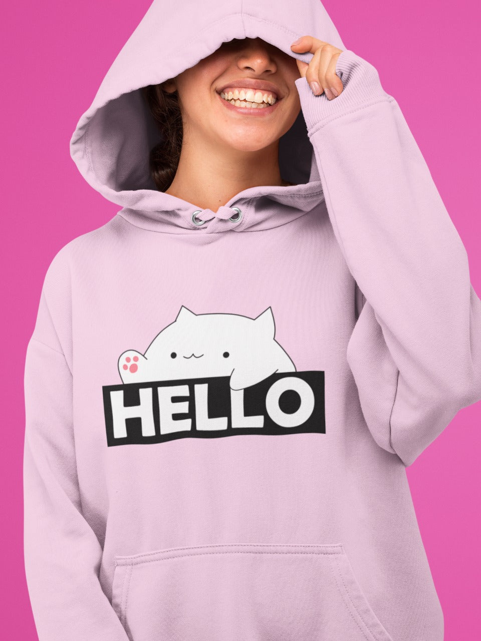 Girl wearing baby pink hoodie with bongo cat saying hello printed on it, the girl's face is half covered with the hood and she is smiling, cute funny memes